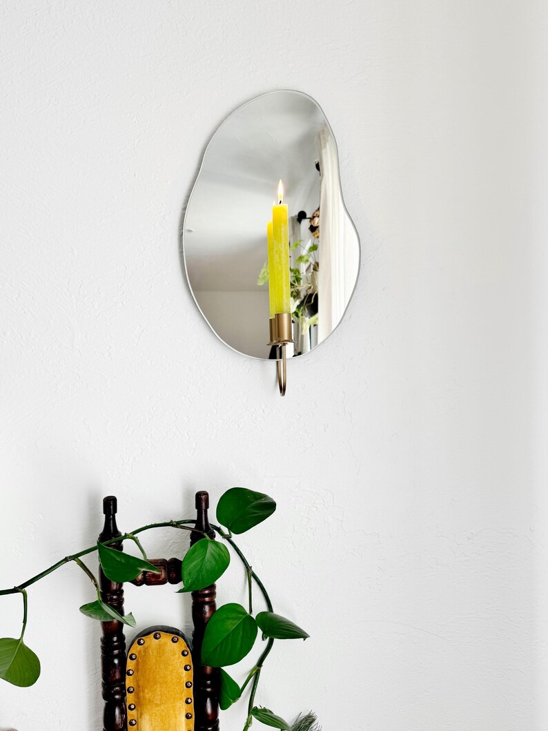 the wall mounted organic shape Mar Mirror Candle Sconce or Mirror Candle Holder. The candleholder is gold & holding a green lit candle. The reflection is of the gold candle holder, the candle & the cozy home setting with plants and decor.