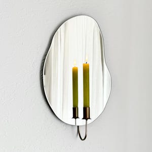 Angled shot of the wall mounted organic shape Mar Mirror Candle Sconce or Mirror Candle Holder. The candleholder is gold & holding a green lit candle. The reflection is of the gold candle holder, the green lit candle & the cream background.