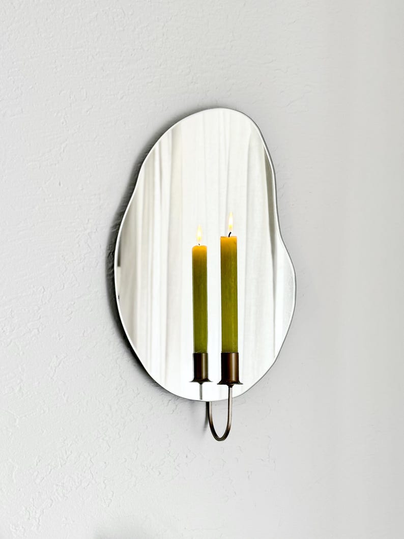 Angled shot of the wall mounted organic shape Mar Mirror Candle Sconce or Mirror Candle Holder. The candleholder is gold & holding a green lit candle. The reflection is of the gold candle holder, the green lit candle & the cream background.