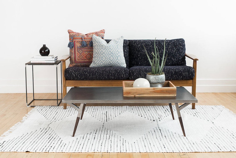 May include: A dark gray couch with wooden arms and legs, two patterned pillows, and a potted plant on a wooden coffee table with a gray top. The couch and coffee table are on a white and black patterned rug.