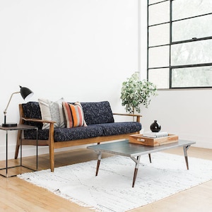 May include: A blue sofa with wooden arms and legs, three pillows, and a black lamp on a black side table. A silver coffee table with a wooden tray on top sits in front of the sofa. A white rug is on the floor.