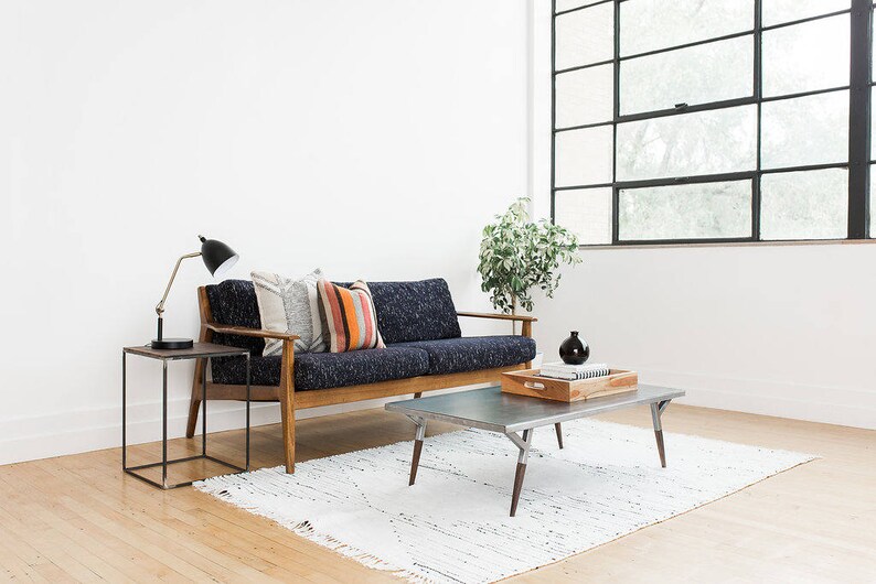 May include: A blue sofa with wooden arms and legs, three pillows, and a black lamp on a black side table. A silver coffee table with a wooden tray on top sits in front of the sofa. A white rug is on the floor.