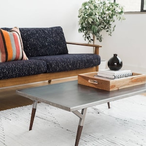 May include: A modern living room with a dark blue sofa, a striped throw pillow, a metal coffee table with wooden legs, a white rug, and a potted plant. The coffee table has a wooden tray with books and a black vase.