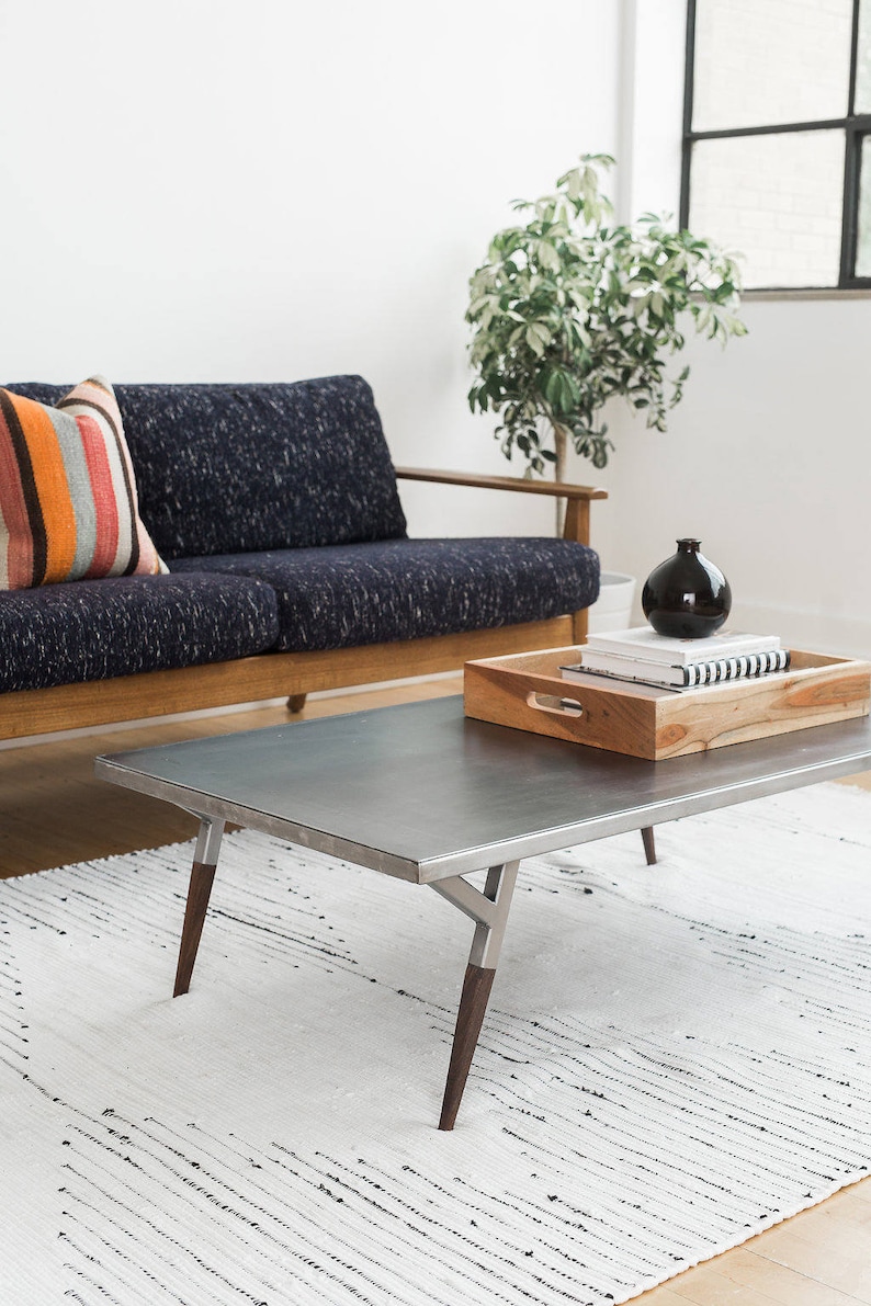 May include: A modern living room with a dark blue sofa, a striped throw pillow, a metal coffee table with wooden legs, a white rug, and a potted plant. The coffee table has a wooden tray with books and a black vase.