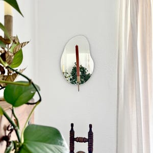 The wall mounted organic shape Mirror Candle Sconce or Mirror Candle Holder. Candleholder is gold, with a red unlit candle. Reflection is: the gold candle holder, the candle & hanging plants. Setting is a cozy home with a vintage chair & plants.