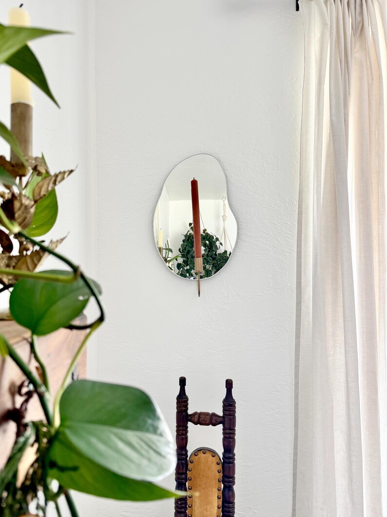 The wall mounted organic shape Mirror Candle Sconce or Mirror Candle Holder. Candleholder is gold, with a red unlit candle. Reflection is: the gold candle holder, the candle & hanging plants. Setting is a cozy home with a vintage chair & plants.