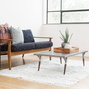 May include: A mid-century modern style sofa with a dark blue fabric upholstery and wooden legs. The sofa has three pillows, one with a red and blue patterned design, one with a white and blue patterned design, and one with a solid blue design. The sofa is in front of a large window with a view of trees. A small wooden coffee table with a metal top sits in front of the sofa. The coffee table has a wooden tray on top with a small potted plant and a white orb.