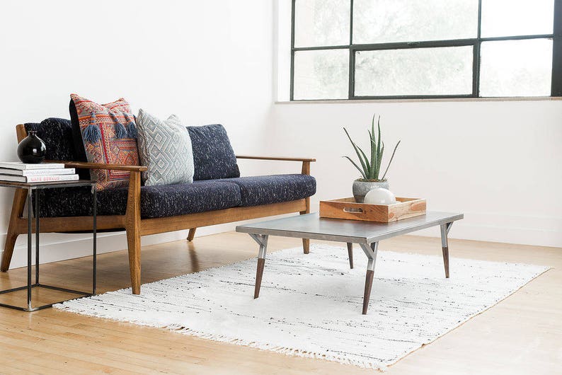 May include: A mid-century modern style sofa with a dark blue fabric upholstery and wooden legs. The sofa has three pillows, one with a red and blue patterned design, one with a white and blue patterned design, and one with a solid blue design. The sofa is in front of a large window with a view of trees. A small wooden coffee table with a metal top sits in front of the sofa. The coffee table has a wooden tray on top with a small potted plant and a white orb.