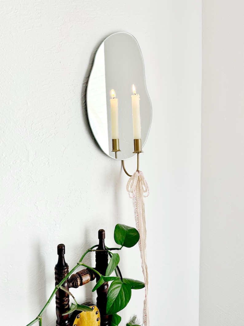 Side angle of the wall mounted organic shape mirror candle sconce or mirror candle holder. The candleholder is gold with lacy ribbon tied to the base to give a whimsical aesthetic. The reflection is of the cream lit candle and candleholder.
