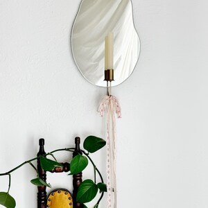 Straight shot of the wall mounted organic shape Mar Mirror Candle Sconce or Mirror Candle Holder. The candleholder has pink & cream ribbons tied in a bow. The candle holder is gold, holding a cream unlit candle. It is a home setting with plants.