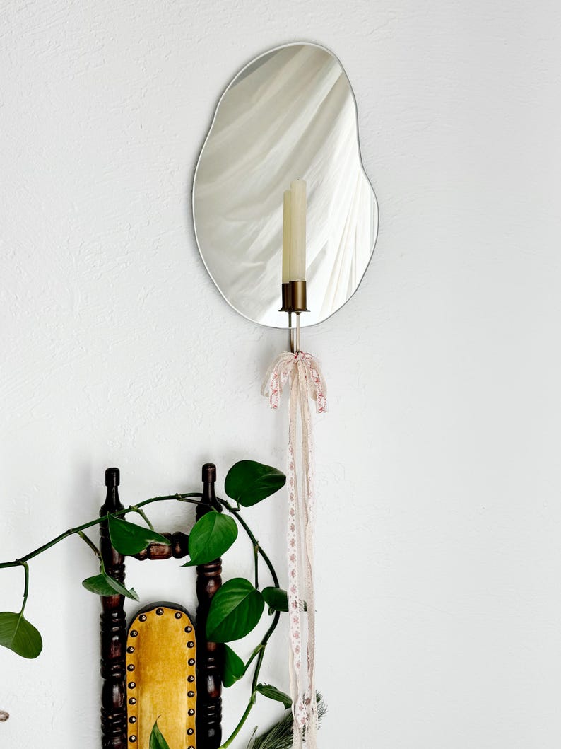 Straight shot of the wall mounted organic shape Mar Mirror Candle Sconce or Mirror Candle Holder. The candleholder has pink & cream ribbons tied in a bow. The candle holder is gold, holding a cream unlit candle. It is a home setting with plants.