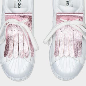May include: A pair of white sneakers with a pink metallic fringe detail over the laces. The shoes have white laces and a white rubber toe cap. The fringe adds a stylish accent to the classic sneaker design.