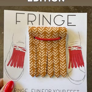 May include: A card with the text "FRINGE" and "LIMITED EDITION" features a mustard yellow and white chevron patterned shoe fringe with a red elastic band. The card also has a drawing of a shoe with red fringe and the text "FRINGE FUN FOR YOUR FEET".