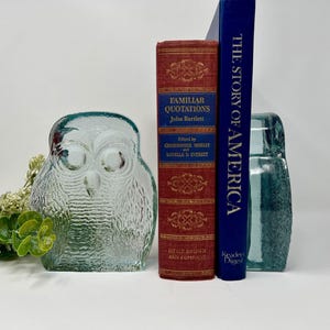 Puede incluir: Una figura de búho de cristal transparente con detalles texturizados se encuentra junto a dos libros. Un libro es rojo con letras doradas, titulado "FAMILIAR QUOTATIONS". El otro libro es azul, titulado "THE STORY OF AMERICA".