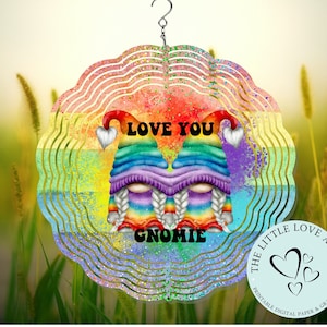 May include: A rainbow-colored wind spinner featuring two gnomes with rainbow hats and braids. The text "LOVE YOU GNOMIE" is displayed. The spinner has a scalloped edge and a silver hanging hook. The background is a blurred image of green grass.