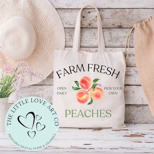 May include: A white canvas tote bag with the text "FARM FRESH" in black lettering. The bag also features a watercolor illustration of peaches with green leaves and the text "OPEN DAILY" and "PICK YOUR OWN" in black lettering. The bag has the text "PEACHES" in green lettering. The bag is sitting on a light wood surface.