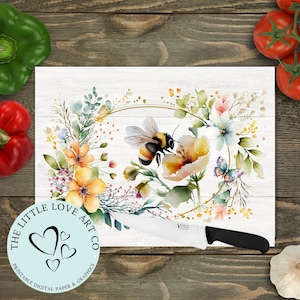 Op de afbeelding: Een wit snijplank met een aquarel bloemenkrans ontwerp met een bij en een vlinder. De krans is omgeven door een gouden cirkel. De tekst "The Little Love Art Co. Printable Digital Paper & Graphics" staat in een cirkel in de linkerbenedenhoek van het bord.