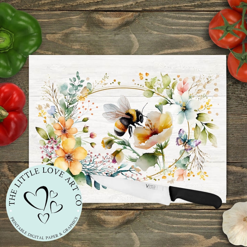 Op de afbeelding: Een wit snijplank met een aquarel bloemenkrans ontwerp met een bij en een vlinder. De krans is omgeven door een gouden cirkel. De tekst "The Little Love Art Co. Printable Digital Paper & Graphics" staat in een cirkel in de linkerbenedenhoek van het bord.