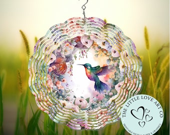 Puede incluir: Un molinillo de viento decorativo de metal con un diseño de colibrí y flores en acuarela. El molinillo tiene un borde festoneado y un gancho para colgar. El colibrí está representado con colores vibrantes azules, verdes y rojos, rodeado de flores rosas y blancas.