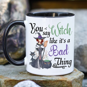 May include: White coffee mug with a black rim and handle. The mug features a cartoon witch with a black cat and a cauldron. The text on the mug reads "You say Witch like it's a Bad Thing."