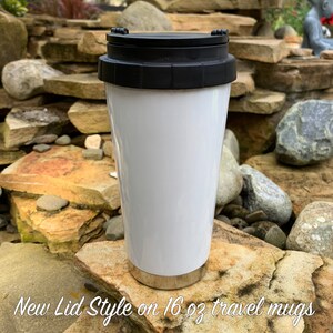 May include: White travel mug with a black lid. The mug is sitting on a rock. The text "New Lid Style on 16 oz travel mugs" is visible below the mug.