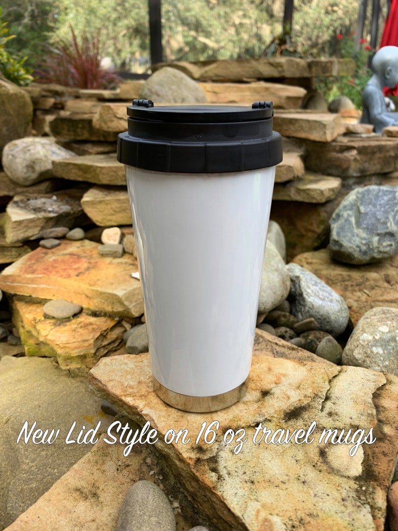 May include: White travel mug with a black lid. The mug is sitting on a rock. The text "New Lid Style on 16 oz travel mugs" is visible below the mug.