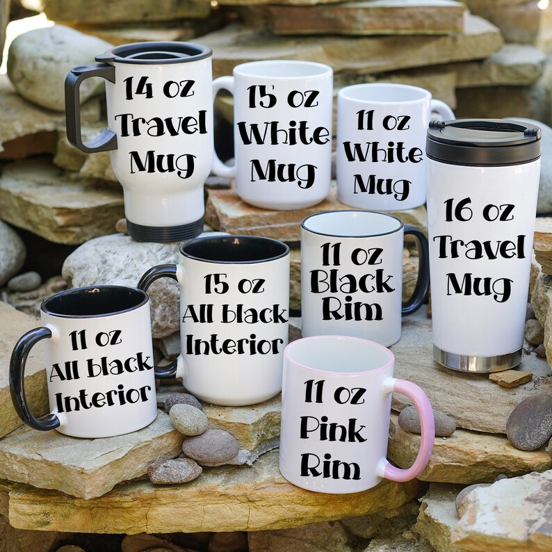 May include: A collection of seven white ceramic mugs with black accents. The mugs are in various sizes, ranging from 11 to 16 ounces. The mugs feature different designs, including a travel mug, a mug with a black rim, and a mug with a pink rim. The mugs are displayed on a background of brown rocks.