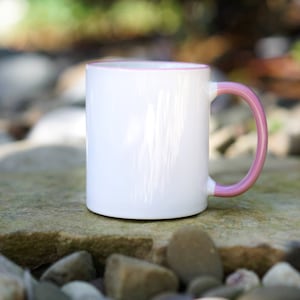 May include: A white ceramic mug with a pink rim and handle. The mug is sitting on a rock surface.