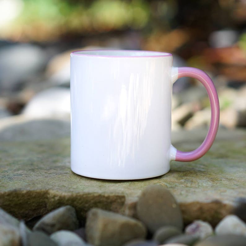 May include: A white ceramic mug with a pink rim and handle. The mug is sitting on a rock surface.