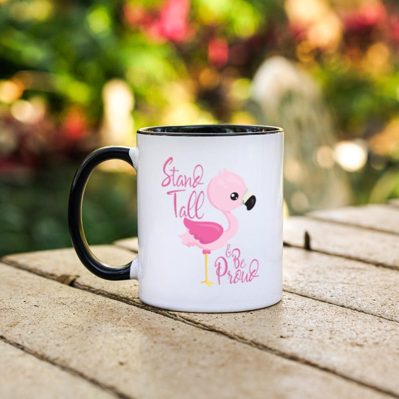 May include: White ceramic mug with a black rim and handle. The mug features a pink flamingo illustration with the text "Stand Tall & Be Proud".