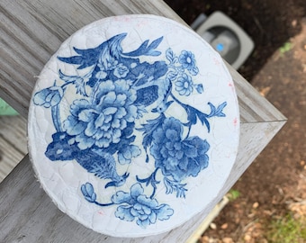 May include: A round, white ceramic coaster with a blue and white floral design. The design features a stylized bird and flowers.