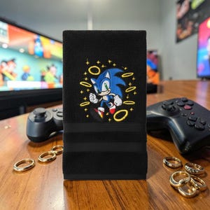 May include: Black hand towel featuring an embroidered image of Sonic the Hedgehog in blue, white, and red. The towel has three black stripes and gold rings around the character. The towel is on a wooden surface.