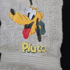 May include: A light gray hand towel with an embroidered image of Pluto, a yellow dog with black ears and a red tongue. The name "Pluto" is embroidered in yellow below the image. The towel has a ribbed border.