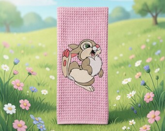 May include: A pink waffle-weave towel featuring an embroidered cartoon rabbit. The rabbit is light brown and white with green eyes and red accents. The towel is displayed on a grassy field with flowers.
