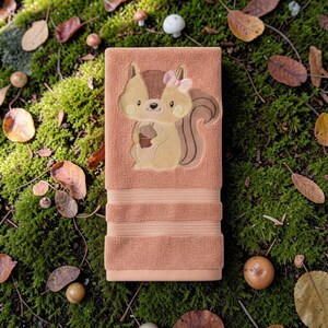 May include: A peach-colored bath towel with an embroidered squirrel design. The squirrel is holding an acorn and wearing a pink bow. The towel has three horizontal stripes near the bottom. The towel is lying on a bed of green moss, leaves, and small mushrooms.