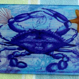 May include: A blue cutting board with a nautical theme. It features a large blue crab, starfish, coral, and seashells. Two orange rectangular sponges are on the right side. The board has a handle with a hole.