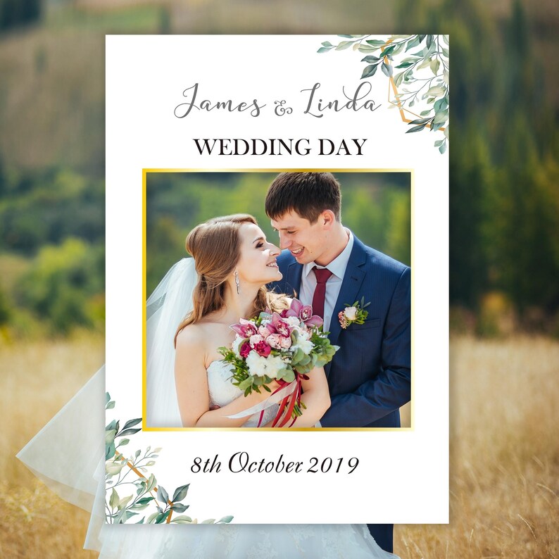 May include: A white wedding photo booth sign with a gold frame and green foliage. The sign says "James & Linda Wedding Day" and "8th October 2019".