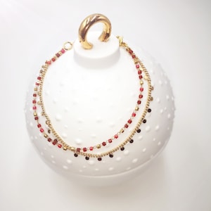 May include: A gold chain anklet with red and gold beads and small gold charms. The anklet is displayed on a white surface.