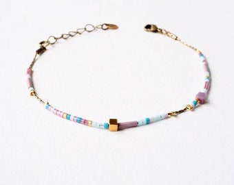 Puede incluir: Un delicado brazalete de cadena dorada con un dije cuadrado y cuentas de colores en tonos rosa, azul y blanco.