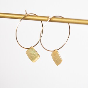 May include: Gold-coloured hoop earrings, each with a hammered, rectangular charm. The earrings are displayed against a white background and a gold-coloured rod. The earrings are a simple, elegant design.