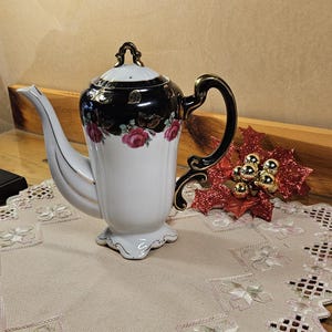 May include: A white and black porcelain teapot with a floral design and gold accents. The teapot sits on a beige embroidered table runner with a floral pattern. A red and gold decorative ornament is to the right.