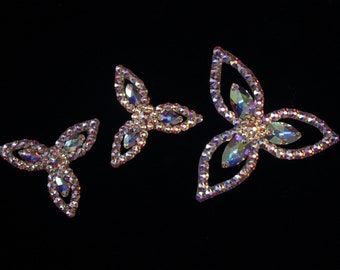May include: Three iridescent rhinestone flower-shaped embellishments on a black background. Each flower has three petals and a central gemstone. The rhinestones reflect a rainbow of colors.