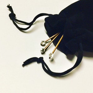 May include: A black velvet drawstring bag with three gold-toned hair pins. Each pin has a clear, round crystal at the top. The bag's drawstring is black, and the background is a light color.