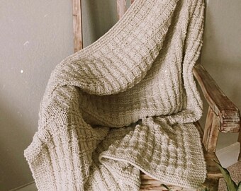 May include: A cream-colored knitted throw blanket draped over a wooden chair. The blanket has a textured, ribbed pattern and is folded over the back of the chair.