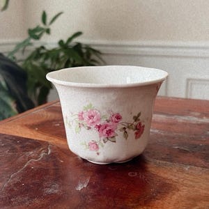 May include: A cream-colored ceramic bowl featuring a speckled texture and a delicate floral pattern of pink roses with green leaves. The bowl has a wide, flared rim and a rounded base, showcasing a soft, light-toned finish with detailed botanical art.