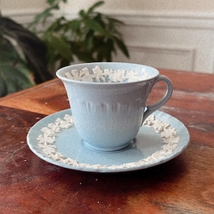 May include: A light blue ceramic teacup and matching saucer set featuring white raised relief floral and vine detailing around the rim of the cup and the edge of the saucer. The teacup has a curved handle and a smooth, glossy finish.
