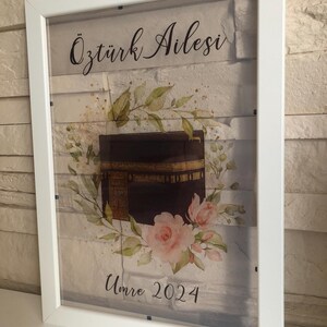 Puede incluir: Una impresi&oacute;n enmarcada en blanco con una ilustraci&oacute;n de acuarela de la Kaaba, un dise&ntilde;o floral y el texto "&Ouml;zt&uuml;rk Ailesi Umre 2024".