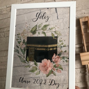 Puede incluir: Una impresi&oacute;n enmarcada en blanco con una ilustraci&oacute;n en negro y dorado de la Kaaba, rodeada de flores de acuarela rosa y verde. El texto "YeliZ Umre 2022 Dez." est&aacute; impreso debajo de la ilustraci&oacute;n.