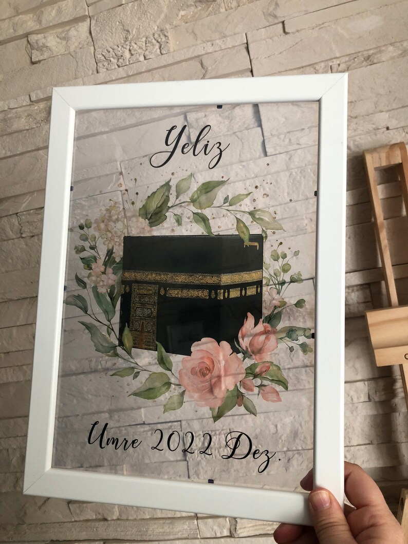 Puede incluir: Una impresi&oacute;n enmarcada en blanco con una ilustraci&oacute;n en negro y dorado de la Kaaba, rodeada de flores de acuarela rosa y verde. El texto "YeliZ Umre 2022 Dez." est&aacute; impreso debajo de la ilustraci&oacute;n.