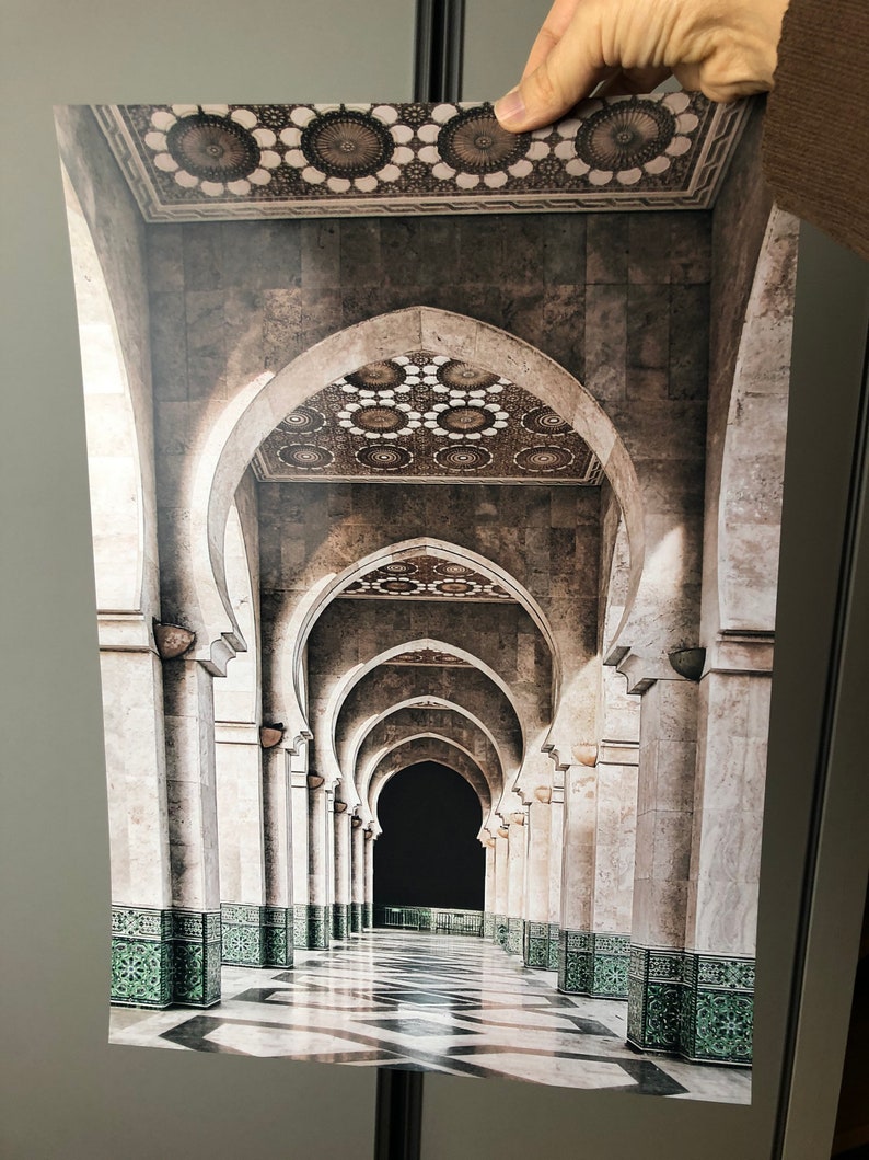 May include: A long hallway with a series of arches and columns. The floor is tiled in black and white, and the walls are a light beige. The arches are decorated with intricate patterns, and the ceiling is adorned with a repeating pattern of circles.